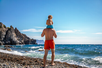 A father carries his little son on his shoulders along the seashore.