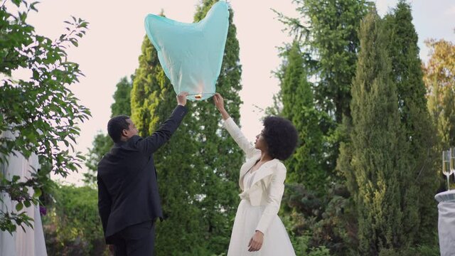 Side view of African American bride and groom raising hands holding chinese lantern together. Young married couple in wedding dress and suit setting off burning air balloon on marriage ceremony