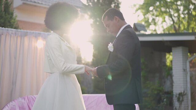 Young loving African American couple standing in sunlight outdoors holding hands looking at each other with love. Side view portrait of affectionate groom and bride in sunrays in garden