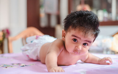 Cute baby girl turning and try to crawl for the first time and looking at the camera, four months old cute daughter with black hair wearing white pampers. Baby photoshoot in 4-month celebration.