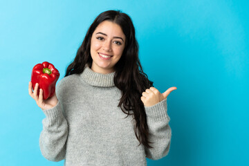 Young caucasian woman holding a pepper isolated on blue background pointing to the side to present a product