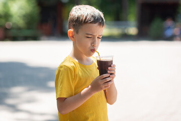a 7-year-old boy in a yellow T-shirt drinks a cool brown drink from a glass to quench his thirst....