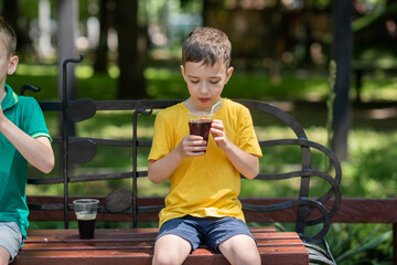 a 7-year-old boy in a yellow T-shirt drinks a cool brown drink from a glass to quench his thirst. Summer heat - obrazy, fototapety, plakaty