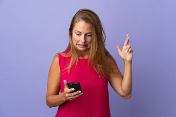 Middle age brazilian woman isolated on purple background using mobile phone with fingers crossing