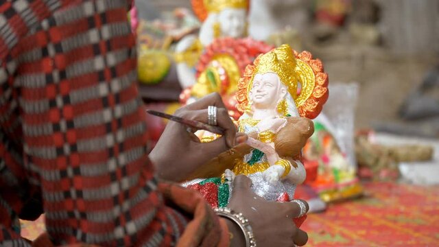 Clay artist putting glitter colors as finishing touches on a small statue of Ma Saraswati. Indian craftsperson coloring the hands of Hindu Goddess for Saraswati puja celebration - an Indian festiva...