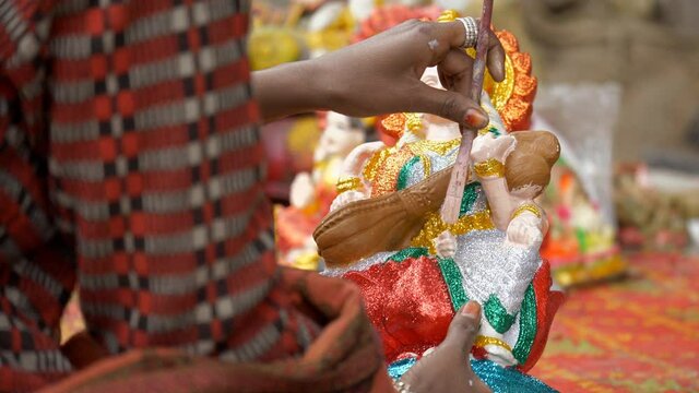 Indian craftsperson giving final touches to a small clay sculpture of Ma Saraswati. Close-up shot of an artist coloring the hands of a beautiful idol/murti of a Hindu goddess for Saraswati puja
