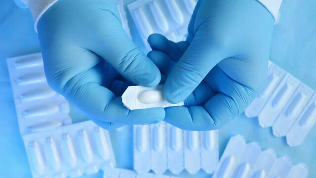 A professional proctologist wearing blue medical gloves opens suppositories for hemorrhoids against the blue backdrop of many other drugs. The Doctor shows candles for the treatment of hemorrhoids