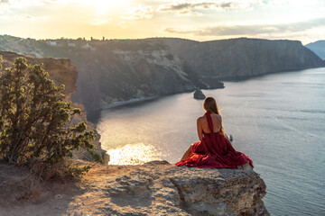 A girl with loose hair in a red dress sits on a rock rock above the sea. In the background, the sun...