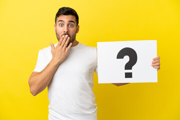 Young handsome caucasian man isolated on yellow background holding a placard with question mark symbol with surprised expression