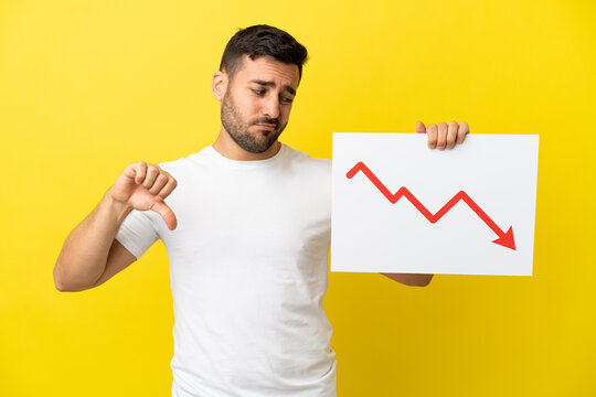 Young handsome caucasian man isolated on yellow background holding a sign with a decreasing statistics arrow symbol and doing bad signal