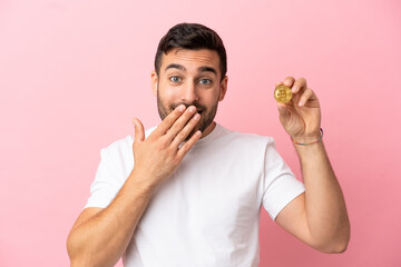 Young man holding a Bitcoin isolated on pink background happy and smiling covering mouth with hand