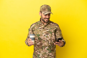 Military man isolated on yellow background holding coffee to take away and a mobile