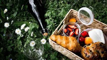 A bottle of cider, picnic basket with fresh croissant, sees and nut bread, strawberry  jam, cherries, cheese on the green grass among daisies, Picnic day outdoors. Close up, macro.