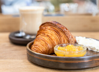 Ceramic plate with fresh crispy croissant, orange jam and butter with a glass cup of cappuccino on wooden table in cafe.