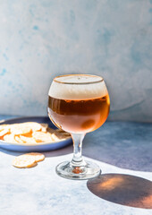 Pint of lager beer in glass with potato crisps, blue concrete background with hard sunlight and shadows. Beer and snack.