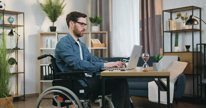 Attractive confident skillful 40-aged bearded handicapped man in wheelchair working on laptop in home office at daytime
