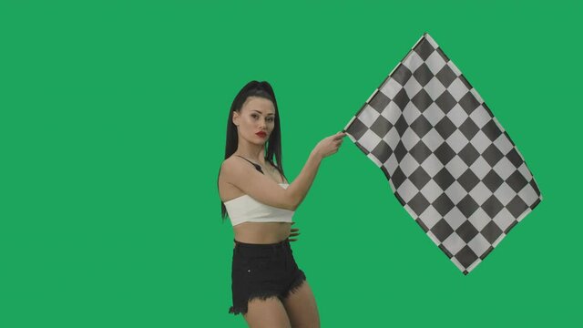 Sexy brunette in shorts waving black and white checkered racing flag to signal start of competition. Young woman posing against background of green screen close up. Slow motion ready, 4K at 59.94fps.
