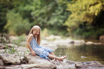 a little girl in the forest on the river sits on a rock splashing water