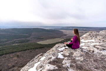 Fototapeta premium A beautiful woman sits in a lotus position on a high place with an amazing view of the mountains and the gorge practicing yoga meditation Kundalini energy thinking intuition prana. Loneliness harmony