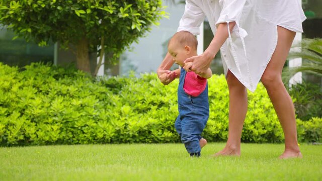 Adorable one year old Baby Girl making her first steps outdoor. Slow Motion Video Footage of the First Steps of the Kid. Sunny day and Green Grass.