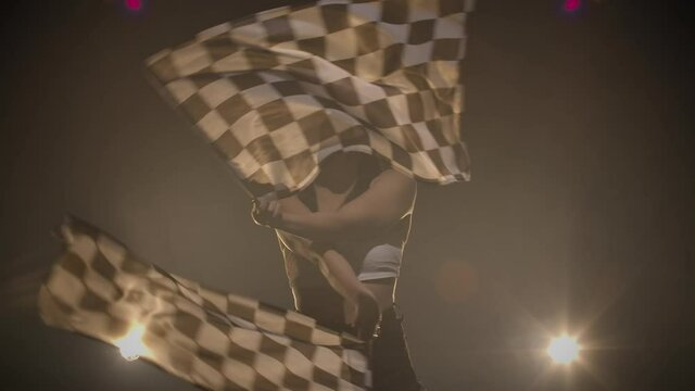 Pretty young woman waving checkered race flags while performing in racing competition. Silhouette of brunette poses in dark smoky studio with backlight. Close up. Slow motion ready, 4K at 59.94fps.
