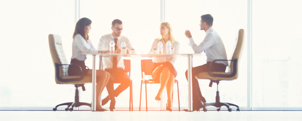 The four business people sit at the office table on the sunny background