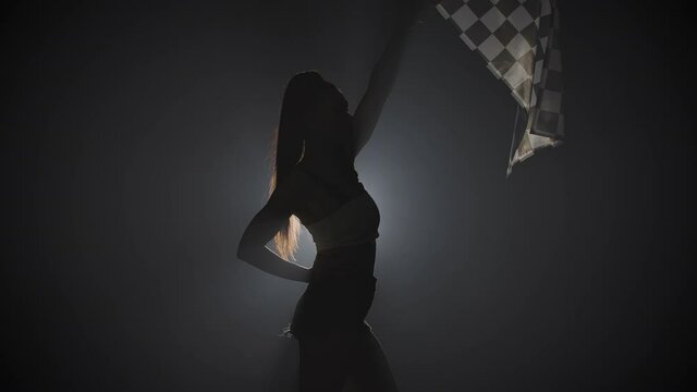 Side view of silhouette young woman waving checkered race flag to signal the start of racing event. Brunette posing in a dark smoky studio with backlight. Close up. Slow motion ready, 4K at 59.94fps.
