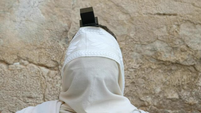 A Jewish man is praying in front of the Wailing Wall, the camera is behind him, focused on the Jewish praying shawl and the Tefillin. a 4K 29.97 fps clip, old city of Jerusalem, Israel.