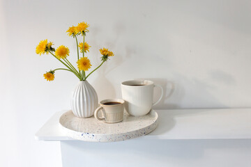 A bouquet of dandelions in a white fluted vase and two cups of different sizes with coffee on a white table.
