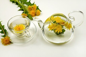 A glass teapot and a cup, with dandelion flower tea in them, stand on a white table.