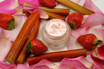 A jar of body care cream stands on the table among the stems of rhubarb, strawberries and rose petals, natural cosmetics