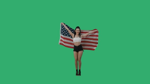 Pretty young woman wrapped herself in flag of united states of america. Smiling brunette posing full length against background of a green screen in the studio. Slow motion ready, 4K at 59.94fps.