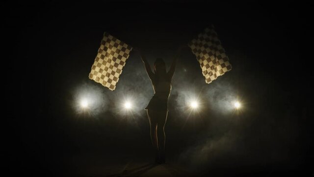 Pretty young woman waving checkered race flags while performing in racing competition. Silhouette of brunette poses full length in a dark smoky studio with backlight. Slow motion ready, 4K at 59.94fps