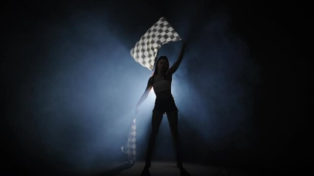 Silhouette of young woman waving a checkered race flag to signal the start of a racing event. Brunette posing full length in a dark smoky studio with backlight. Slow motion ready, 4K at 59.94fps.