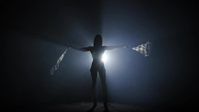 Rear view of silhouette young woman waving checkered race flag to signal the start of racing event. Brunette posing full length in dark smoky studio with backlight. Slow motion ready, 4K at 59.94fps.