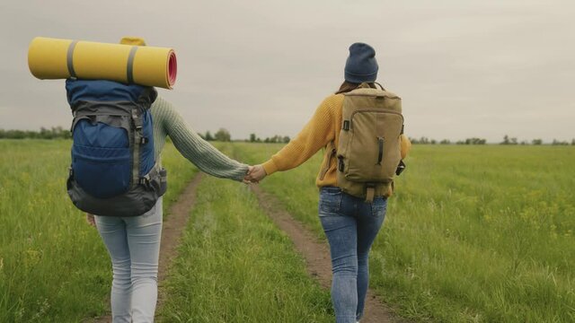 Two hipster girls with backpacks go on a hike holding hands, happy millennial travelers are walking along a dirt road and looking for adventure, time to dream in nature, active people