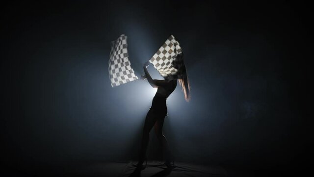 Side view of silhouette young woman waving checkered race flag to signal the start of racing event. Brunette posing full length in dark smoky studio with backlight. Slow motion ready, 4K at 59.94fps.