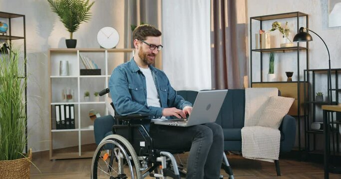 Handsome smiling confident adult bearded disabled man in glasses sitting in wheelchair after accident at home and working on computer ,front view