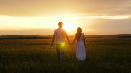 Silhouette of man and a woman at sunset. Romantic date and love in nature. A beautiful girl and a guy are walking hand in hand in field, in sun on the green grass. A young couple in love is traveling.