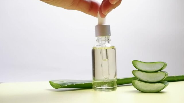 A female hand holds a cap with a pipette from which drops of lotion, serum are dripping into a bottle of aloe vera gel with aloe vera leaves on a white background.