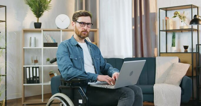 Handsome Concentrated Creative Adult Bearded Disabled Man In Glasses Sitting In Wheelchair Working On Laptop At Home And Looking At Camera With Confident Face Expression