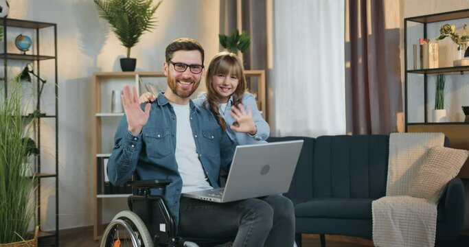 Lovely smiling 10-aged girl hugging her joyful bearded father which sitting in wheelchair after accident and together looking at camera with happy faces and waving hands