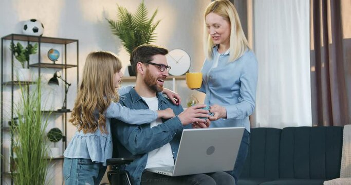 Family idyll where beautiful adult blondie giving two cups of tea to her cute smiling teenage daughter and handsome bearded disabled husband in wheelchair which together applying laptop at home