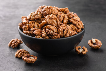 Peeled walnut in a bowl and a few nuts scattered around on a black textured background, side view.