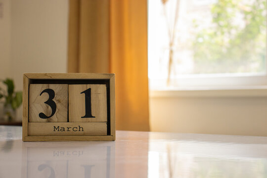 Wooden blocks of the calendar represents the date 31 and the month of March on the background of a window, curtain and a plant.