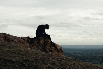 A mental health concept of a hooded man sitting alone on top of a hill in the countryside. Looking...