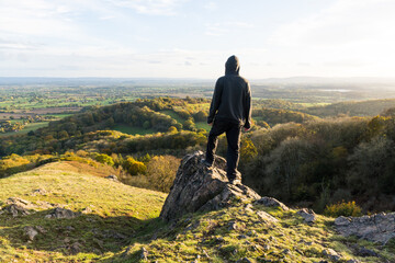 A hooded man, standing on a rocky outcrop on top of a hill, Looking out across the countryside in...
