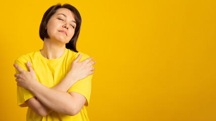 Banner. Young woman wearing casual t-shirt over yellow background, hugging oneself happy and positive, smiling confident. Self love and self care.