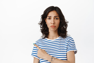 Sad cute brunette woman pointing left, frowning and looking upset, show unfair bad thing, disappointed with advertisement, standing against white background