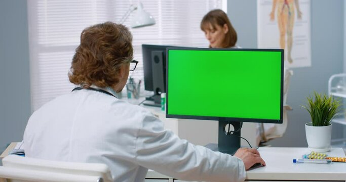 Caucasian doctor sits in hospital office and works at computer typing on keyboard. View from back. Chromakey.